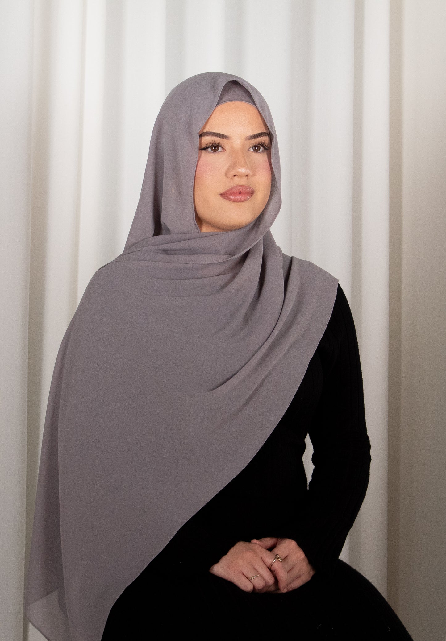Woman wearing a gray hijab and black outfit against a white curtain