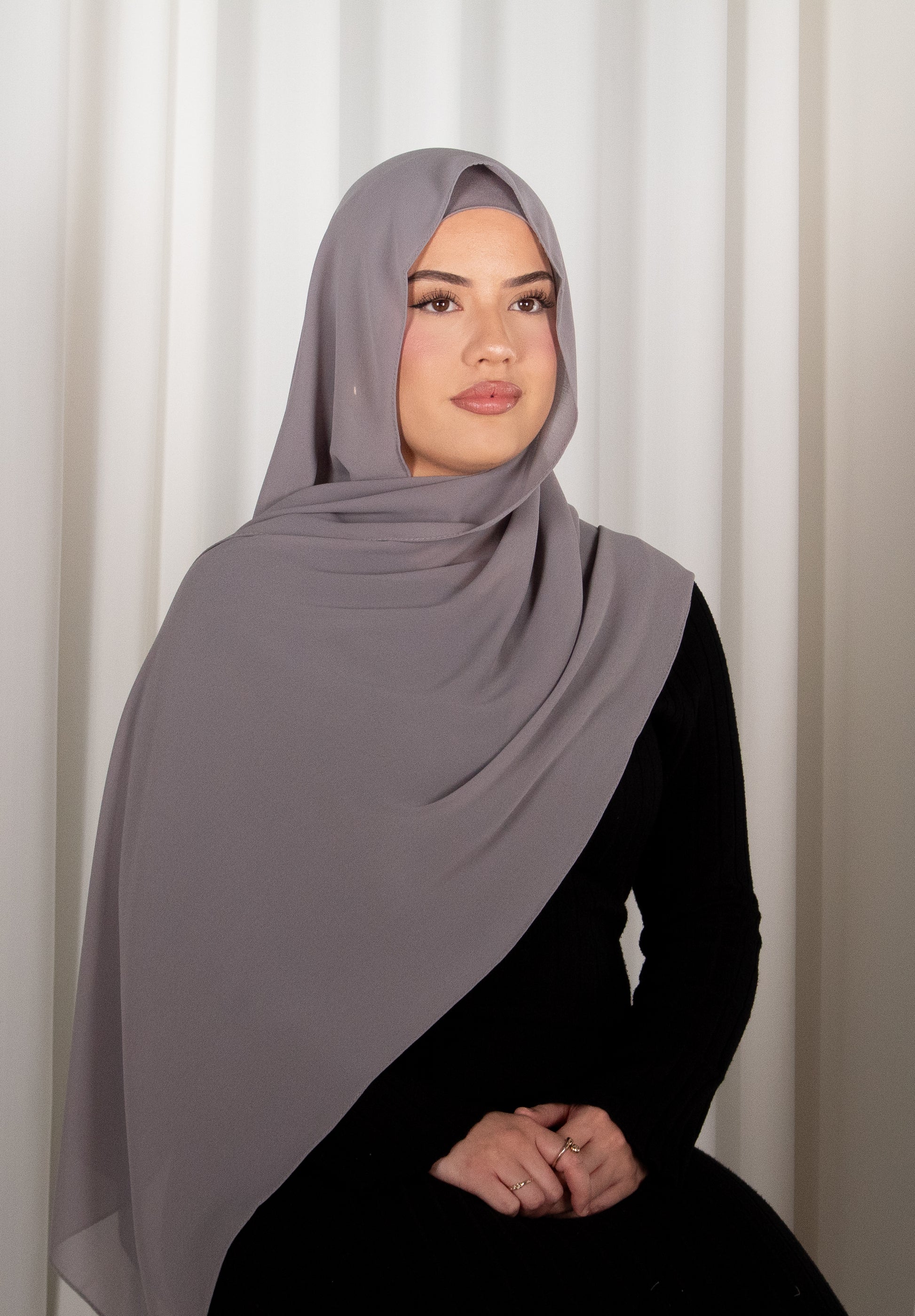 Woman wearing a gray hijab and black outfit against a white curtain