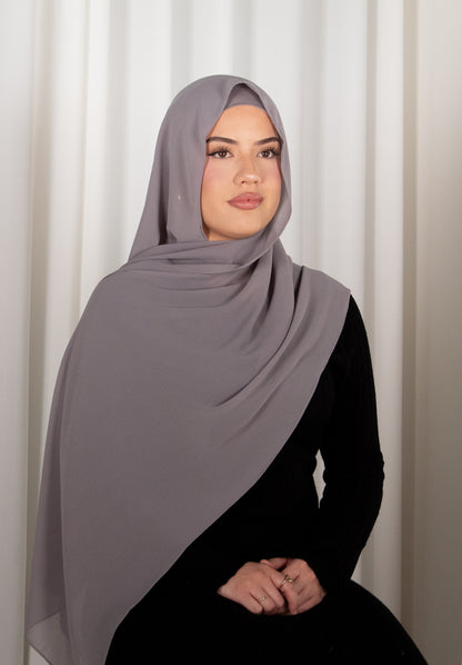 Woman wearing a gray hijab and black outfit against a white curtain