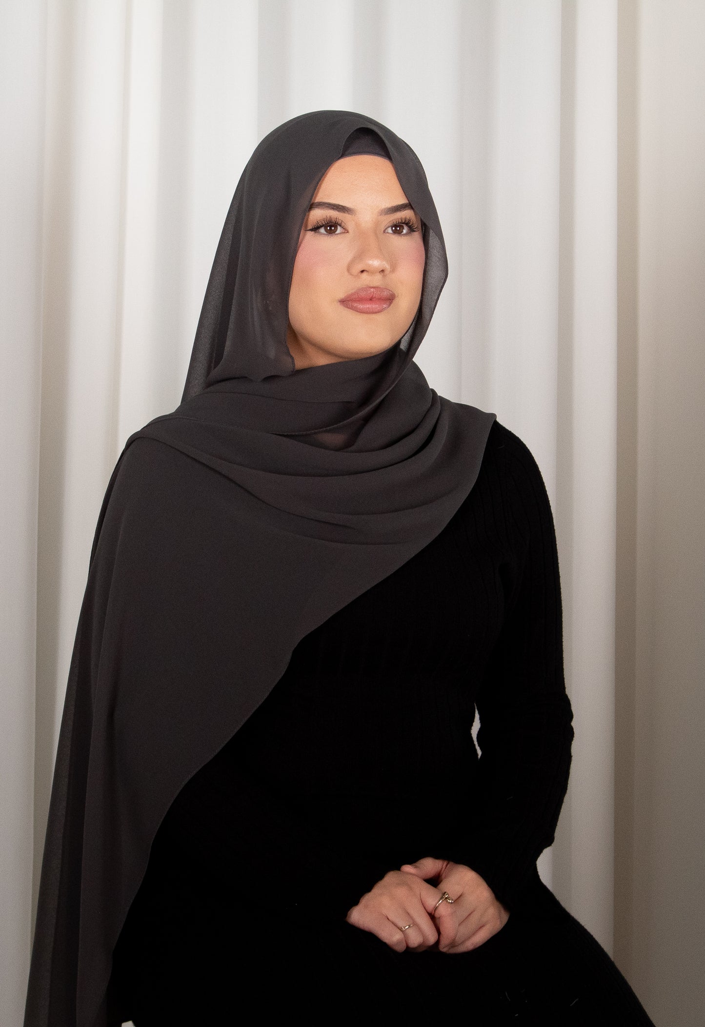 Woman wearing a dark hijab and black outfit against a white curtain background
