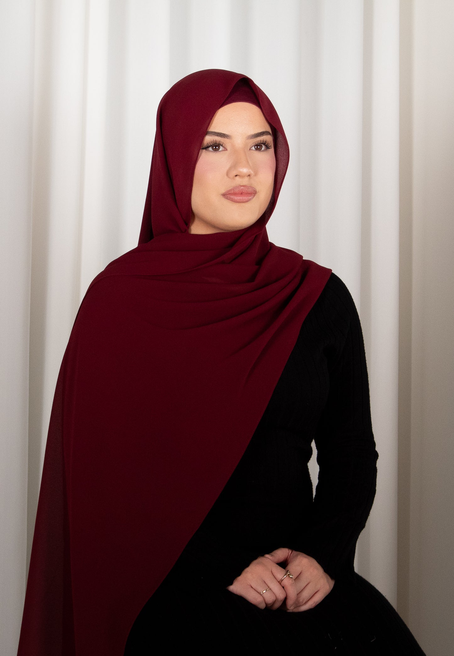 Woman wearing a burgundy hijab and black outfit against a white curtain background