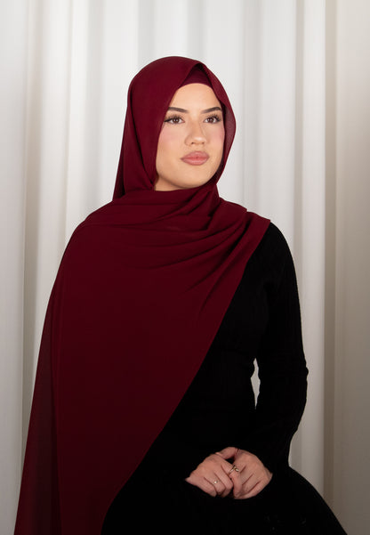 Woman wearing a burgundy hijab and black outfit against a white curtain background
