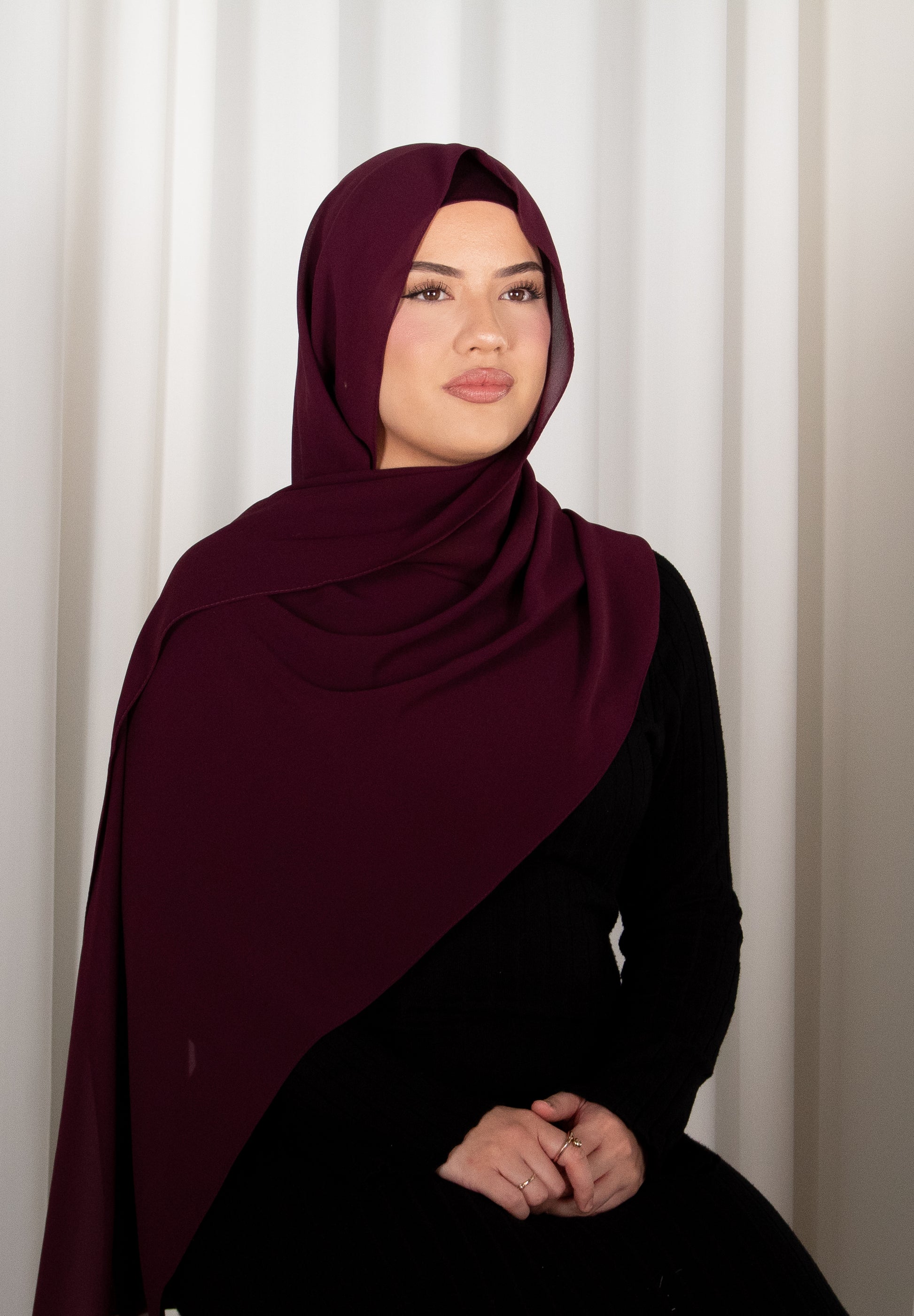 Woman wearing a dark red hijab and black outfit against a white curtain background