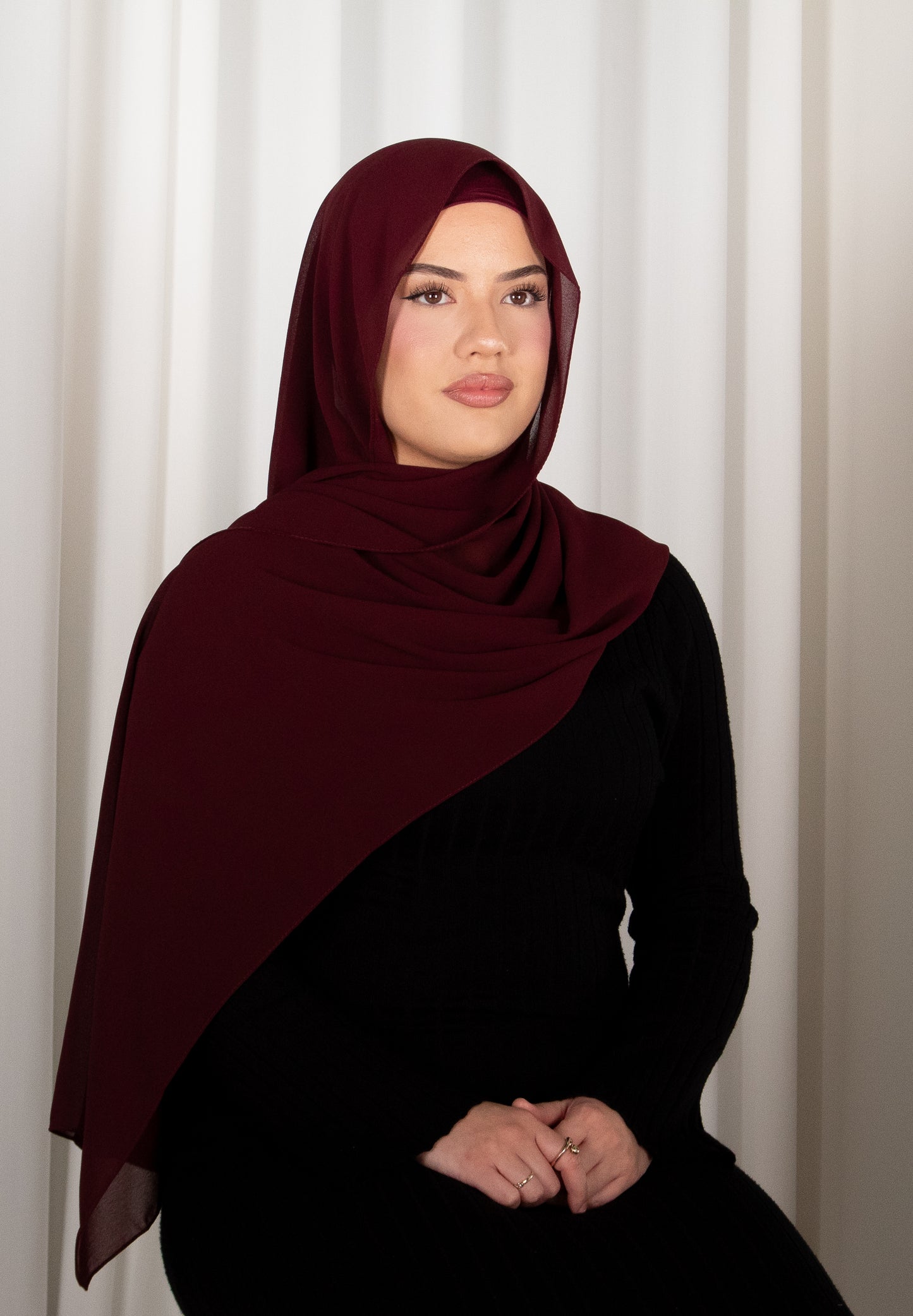 Woman wearing a burgundy hijab and black outfit against a white curtain background