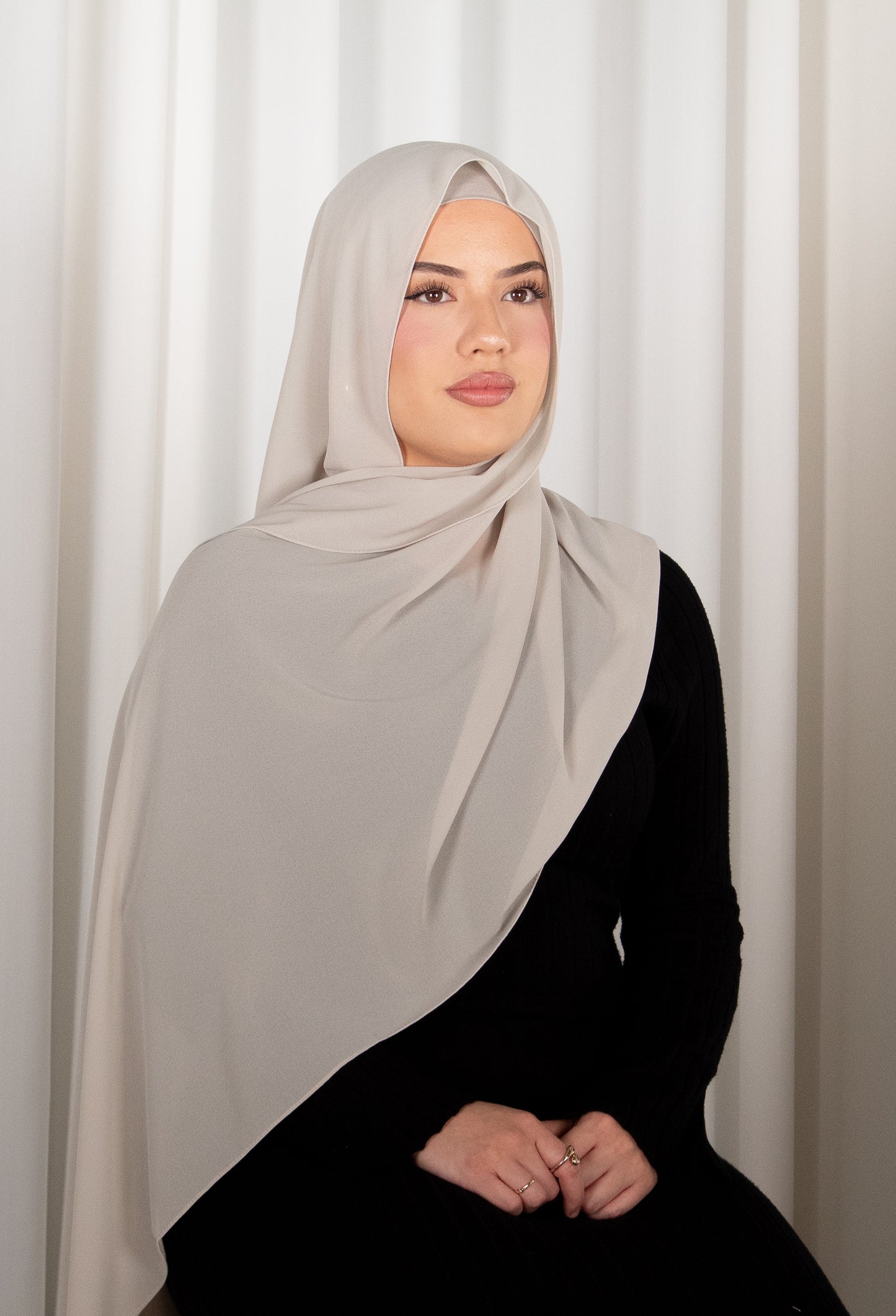 Woman wearing a light gray hijab and black outfit against a white curtain background