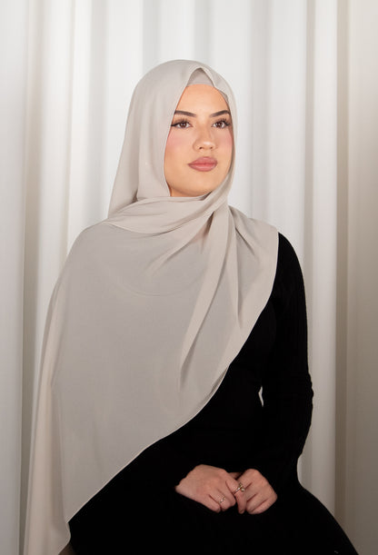 Woman wearing a light gray hijab and black outfit against a white curtain background