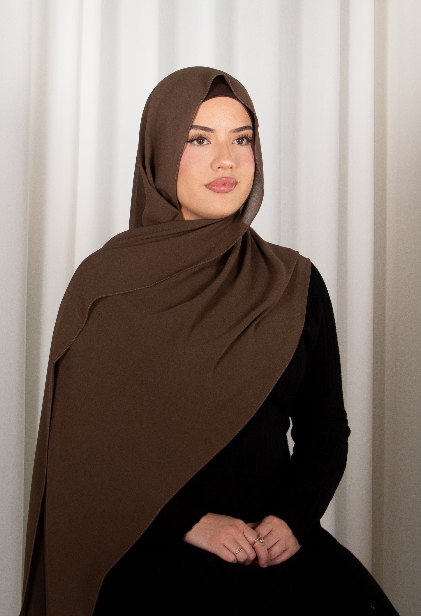 Woman wearing a brown hijab and black outfit against a white curtain background