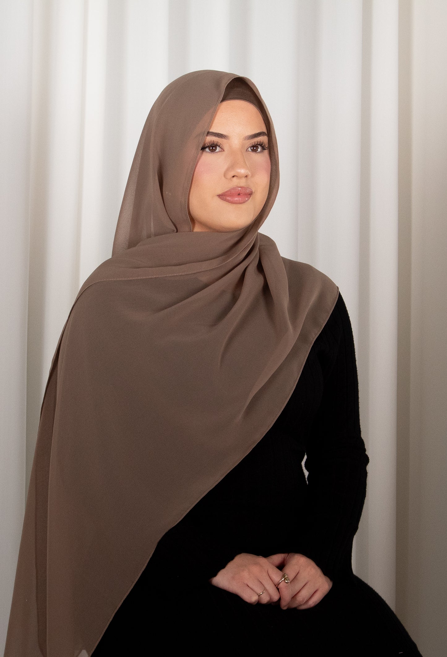 Woman wearing a brown hijab and black outfit against a white curtain background