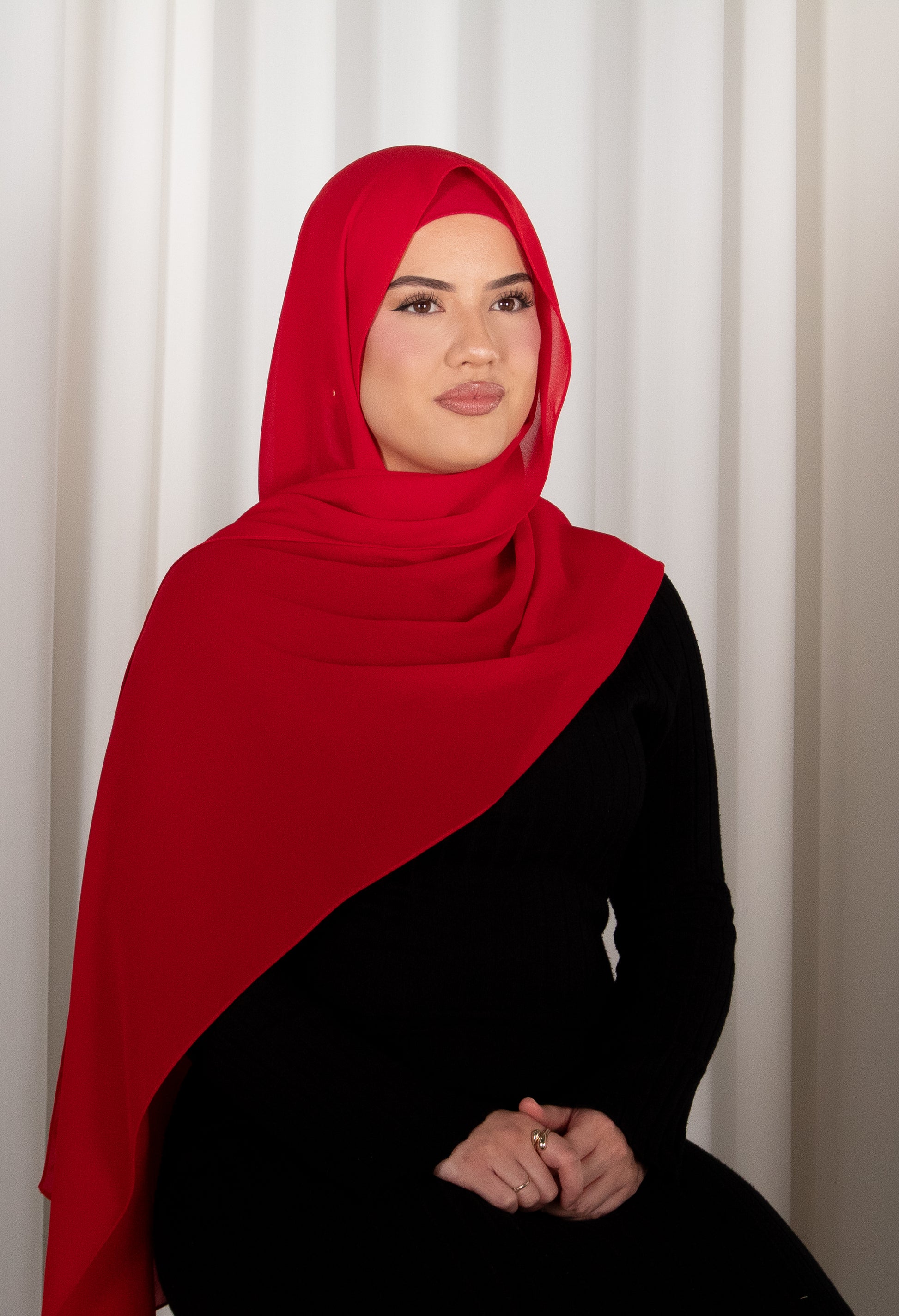 Woman wearing a red hijab and black outfit against a white curtain background