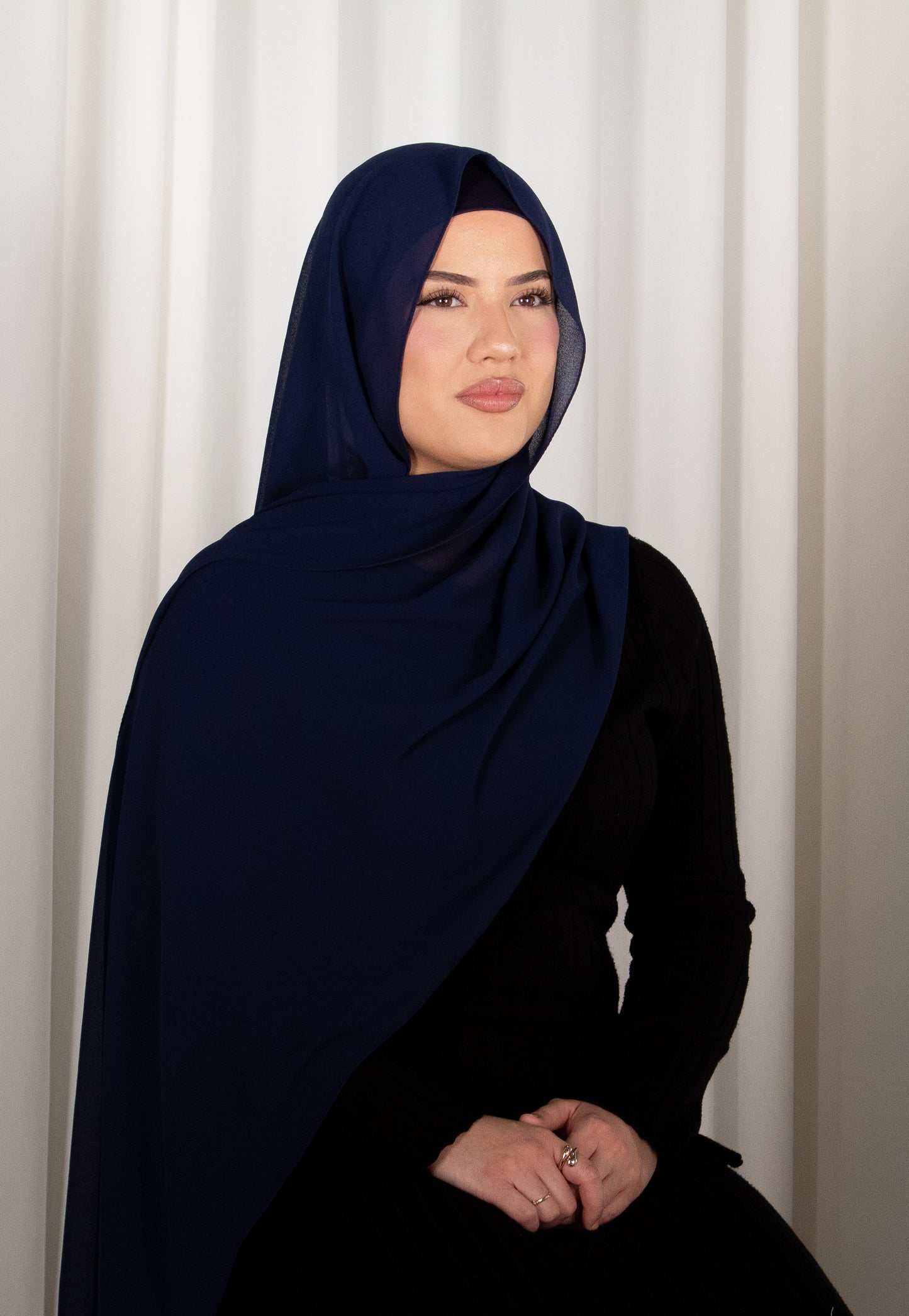 Woman wearing a navy hijab and black outfit against a white curtain background