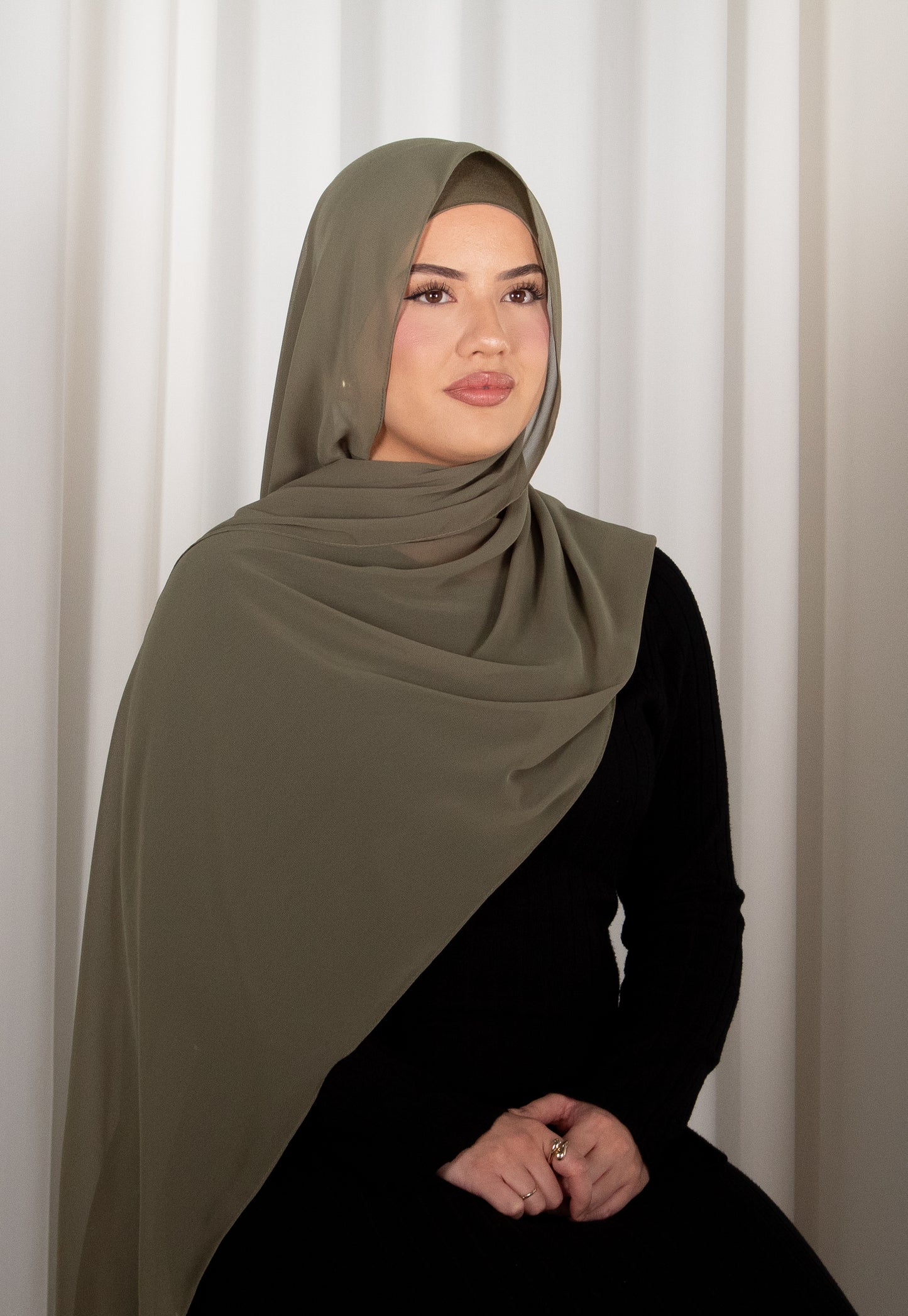 Woman wearing a green hijab and black outfit against a white curtain background