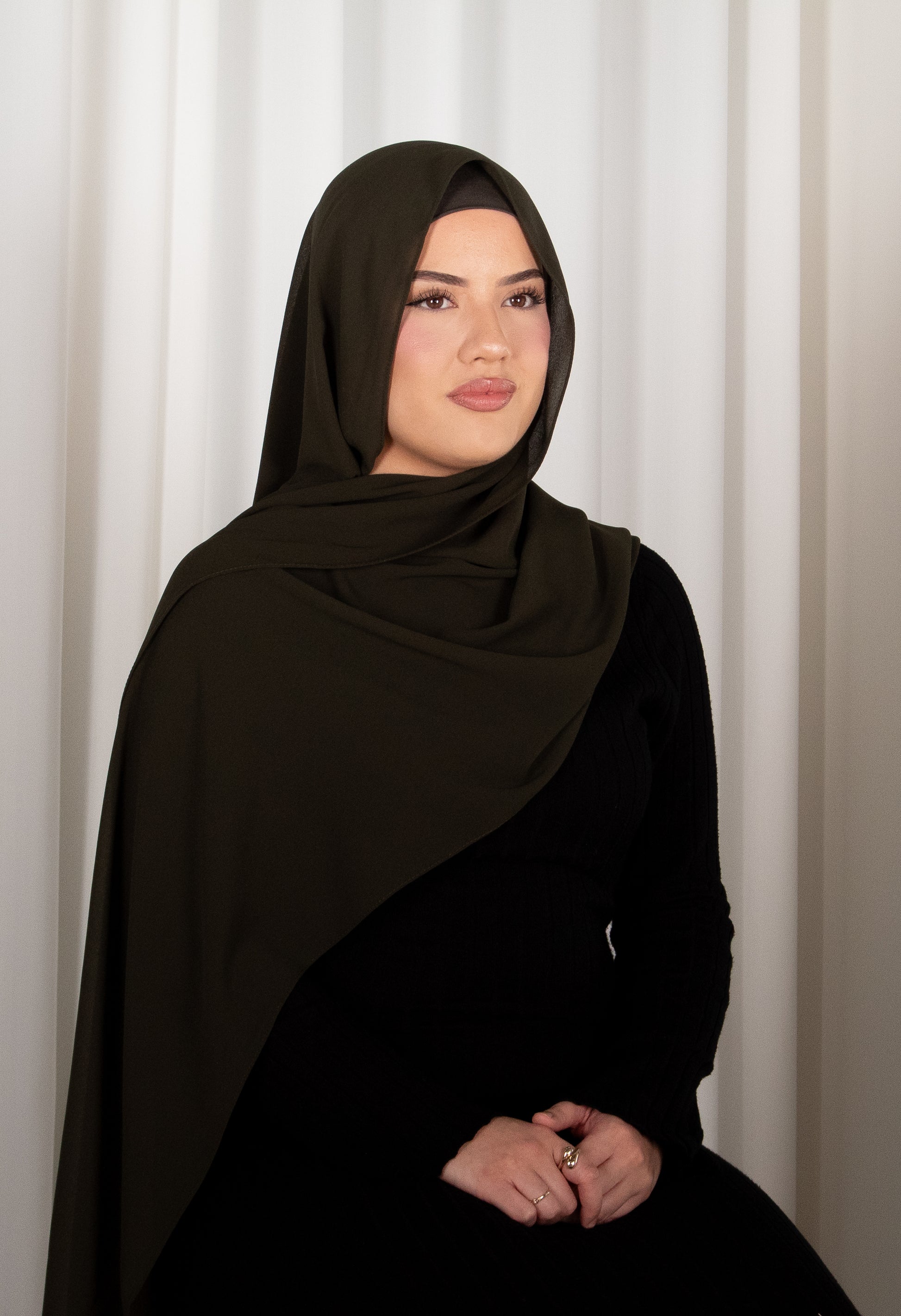 Woman wearing a black hijab and black outfit against a white curtain background