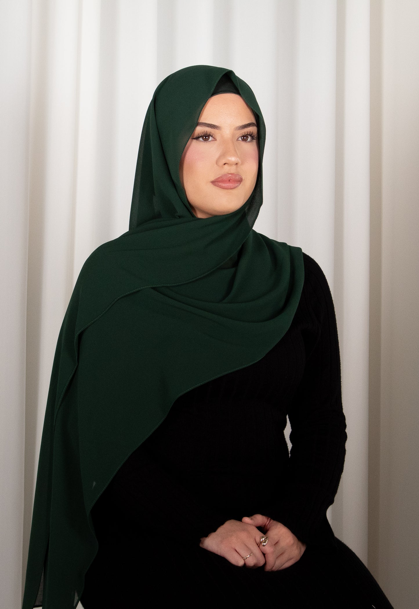 Woman wearing a dark green hijab and black outfit against a white curtain background